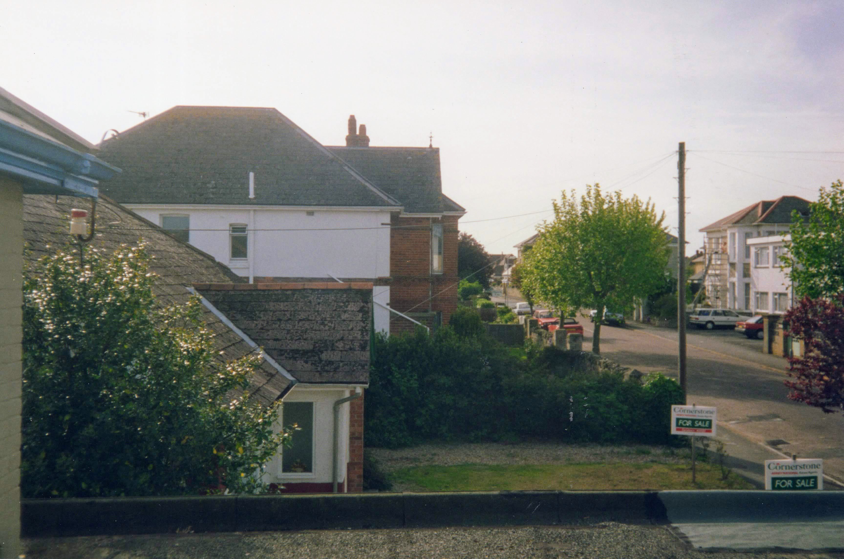 A photograph taken from a hotel bedroom window. In the immediate foreground is the edge of a flat roof. Beyond that, the garden of a neighbouring house with For Sale signs. Next to that, a two-storey house, painted white, with a distinctive decorative line feature. On the other side of the road in the middle distance is a short, flat-roofed, two-storey building.