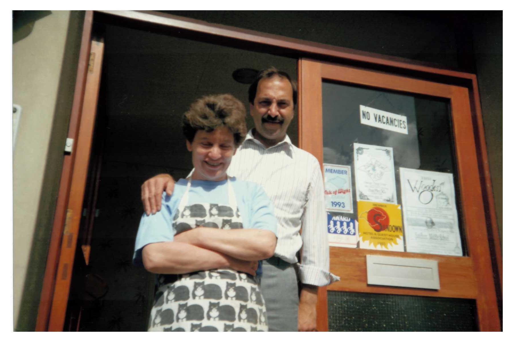 A photo of a female and male couple standing in a doorway. Behind them are various posters and stickers on a window. One of them clearly shows a Sandown tourism message.