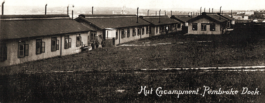 A black and white photograph of six single-storey barrack huts, with Pembroke Dock in the background.